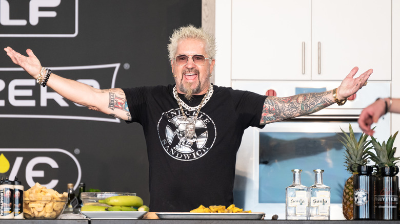 Guy Fieri in a kitchen on sage with arms open