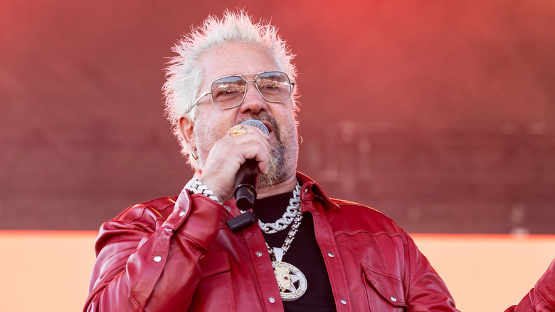 Guy Fieri wearing a red leather jacket and speaking into a microphone.