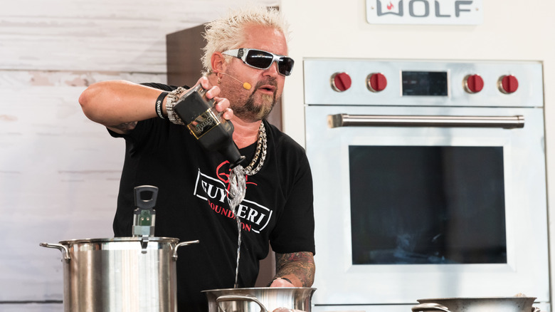 Guy Fieri performing a live cooking demonstration, pouring a liquid into a pot.