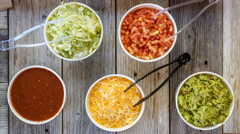 A taco station has ingredients like tomato, cheese, guacamole, and salsa.