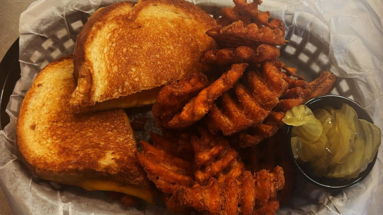 A grilled cheese in a basket with a side of sweet potato waffle fries and pickles at Mac & Ernie's