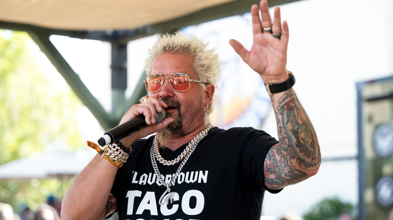 Guy Fieri speaking into a microphone while holding his left hand up