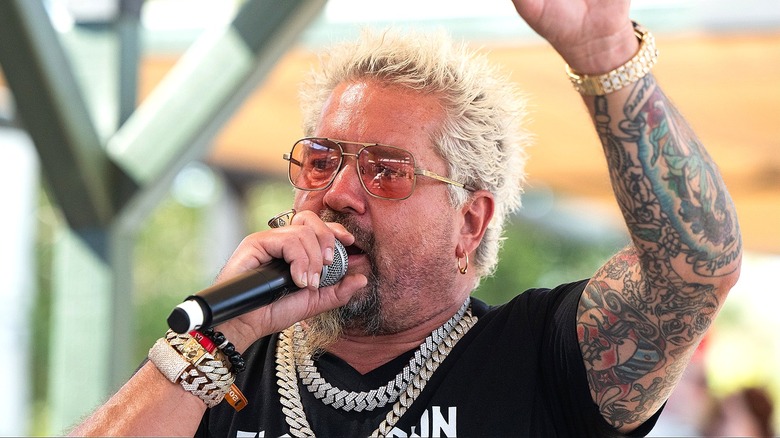 Guy Fieri wearing tinted sunglasses and speaking into a microphone