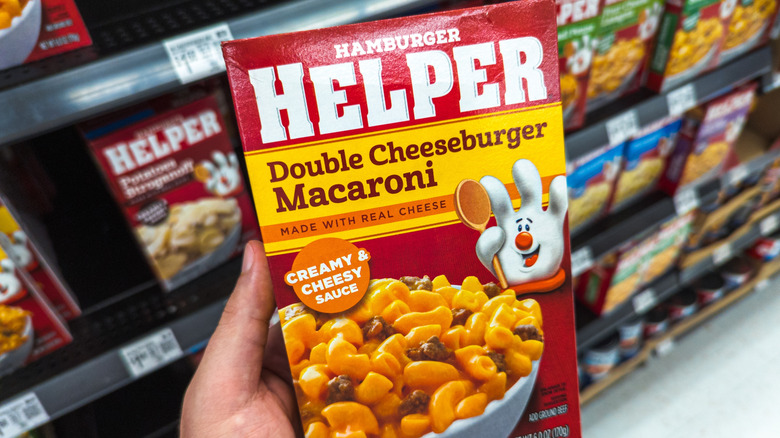 Hand holding a box of Hamburger Helper double cheeseburger macaroni with shelves of Hamburger Helper in background