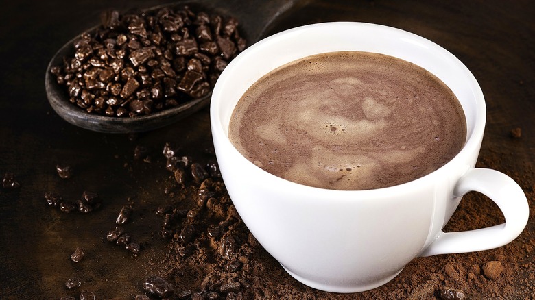Hot chocolate in a white mug with handle on a brown surface covered in cacao nibs and cocoa powder
