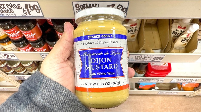 Hand holding a jar of Trader Joe's Dijon mustard in the store's condiment aisle