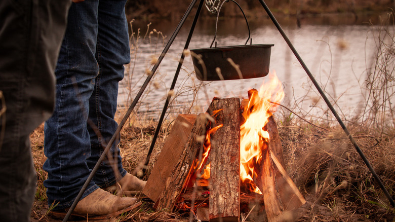 A person wearing cowboy boots standing next to a pot suspended over a wood fire