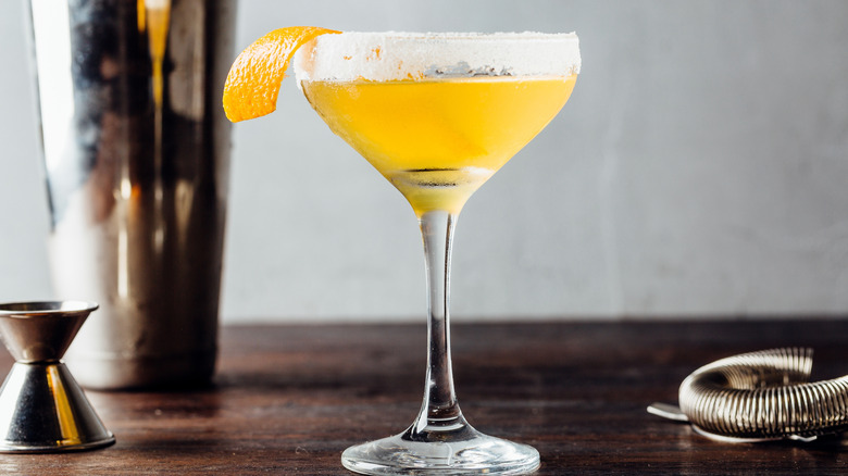 A sidecar cocktail with a lemon twist garnish on a dark wood table next to a stainless steel jigger, cocktail shaker, and strainer.