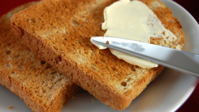 A knife is seen spreading soft butter on toast.