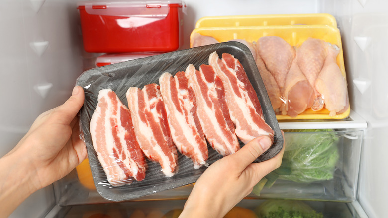Hands holding a package of bacon in front of an open refrigerator. 