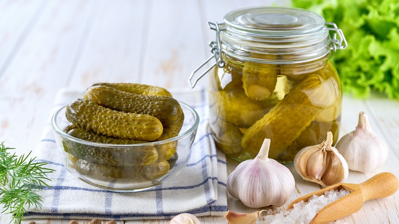 A small bowl of homemade pickled cucumbers next to a jar full of them, surrounded by fresh garlic, dill, and salt.