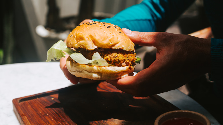 someone's hands holding a vegan burger in a bun with iceberg lettuce over a wooden chopping board