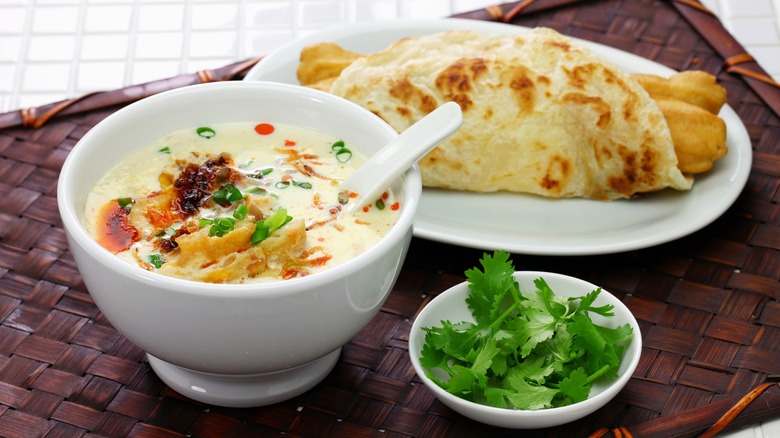 A bowl of savory soy milk next to fried dough sticks.