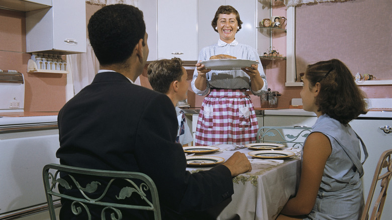 Mother in 60s style kitchen serving dinner to a family at the table.