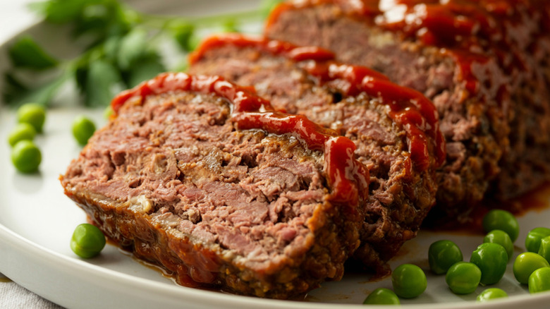 Meatloaf with ketchup glaze on a white plate with peas.