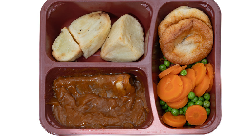 TV dinner with meat, bread, and vegetables in a maroon plastic tray.
