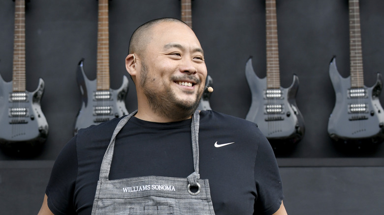 David Chang stands smiling in an apron before a wall of black electric guitars