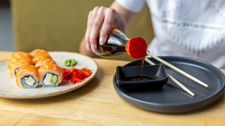 Close-up of soy sauce being poured into a dipping bowl beside salmon sushi rolls, chopsticks, ginger, and wasabi.