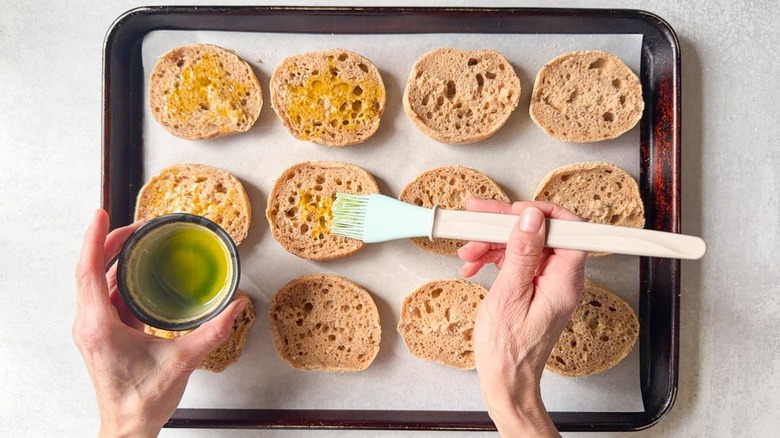 Brushing olive oil on English muffin halves on parchment-lined baking sheet with pastry brush