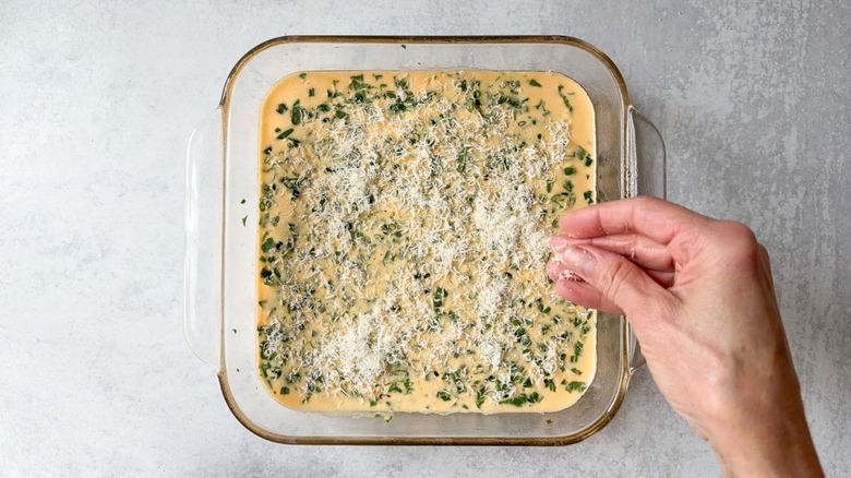Sprinkling grated parmesan onto egg filling mixture in square, glass baking dish