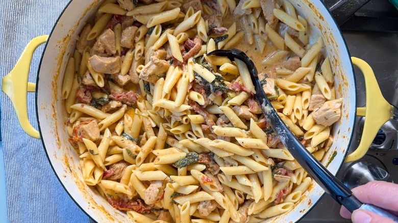 Stirring high protein marry me chicken pasta with pasta spoon in large yellow pot on stovetop