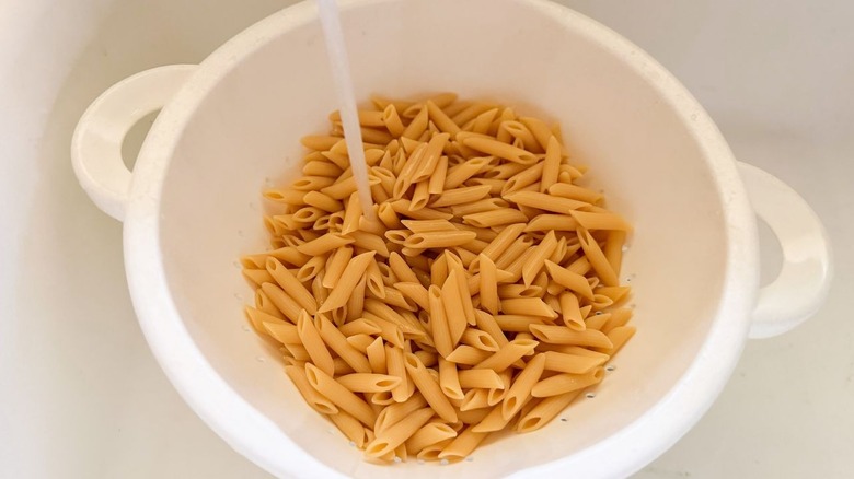 Rinsing penne pasta with water in white colander in sink