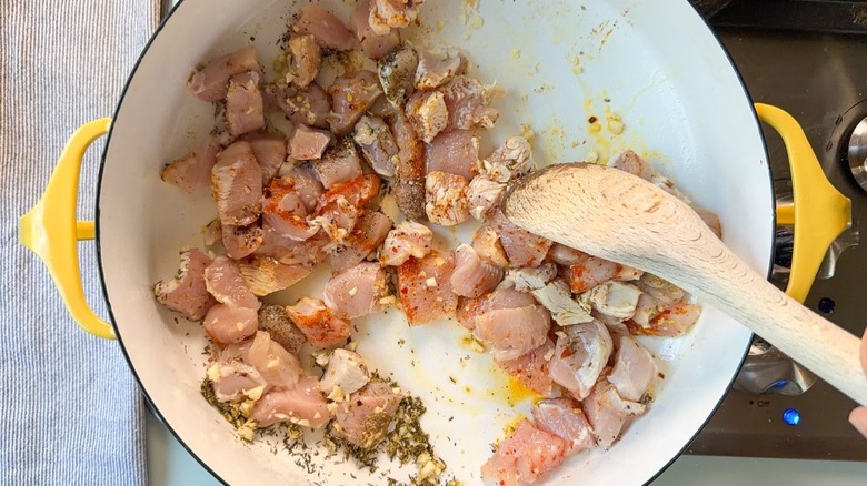 Stirring cubed raw chicken breasts and spices with wooden spoon in large yellow pot on stovetop