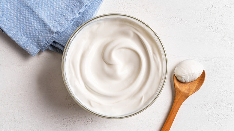 Bowl of plain yogurt next to dishcloth and wooden spoon on white counter.