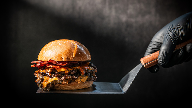 Bacon cheeseburger on a spatula held by a gloved hand