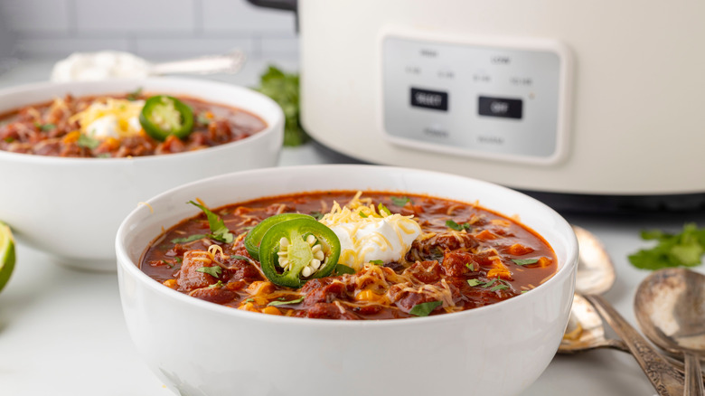 A bowl of chili topped with cheese, sour cream, and jalapeño slices, with a white slow cooker in the background