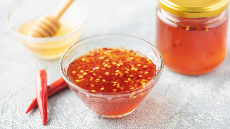 A glass bowl of hot honey, accompanied by two red chili peppers, and in the background a bowl of honey with a wooden honey dipper and a glass jar of honey with a gold lid