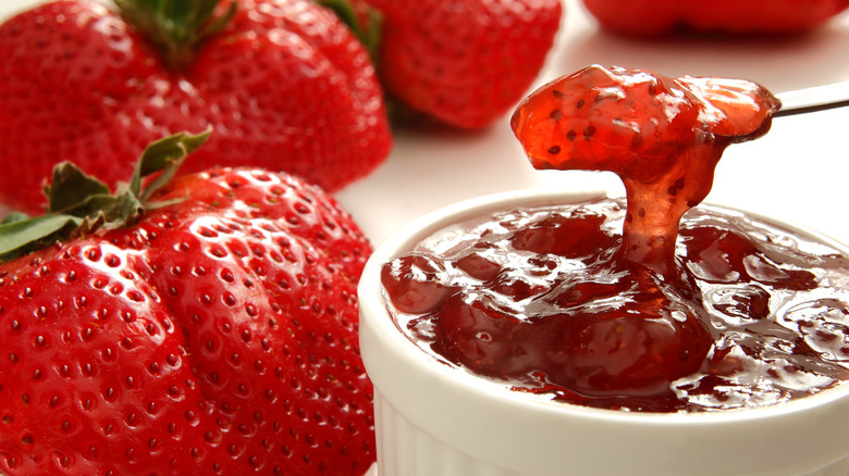 Knife dipping into white ramekin full of strawberry jam, which is sitting next to several strawberries