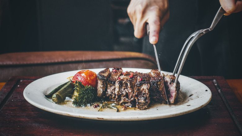 A chef cutting a perfectly cooked ribeye on a plate with a roasted tomato and greens