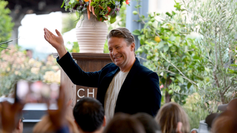A smiling Jamie Oliver with one hand raised in front of an audience