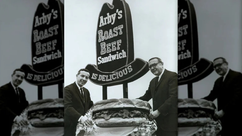 Arby's founders standing with a giant roast beef sandwich in front of an Arby's sign