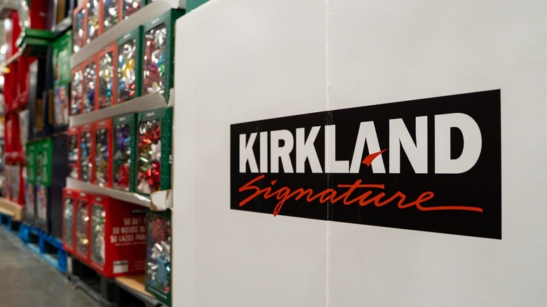 The Kirkland Signature logo inside of a Costco warehouse with boxes of products on shelves behind it.