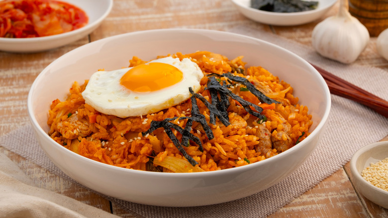 Kimchi fried rice tipped with a fried egg and seaweed, kimchi and whole garlic in the background.