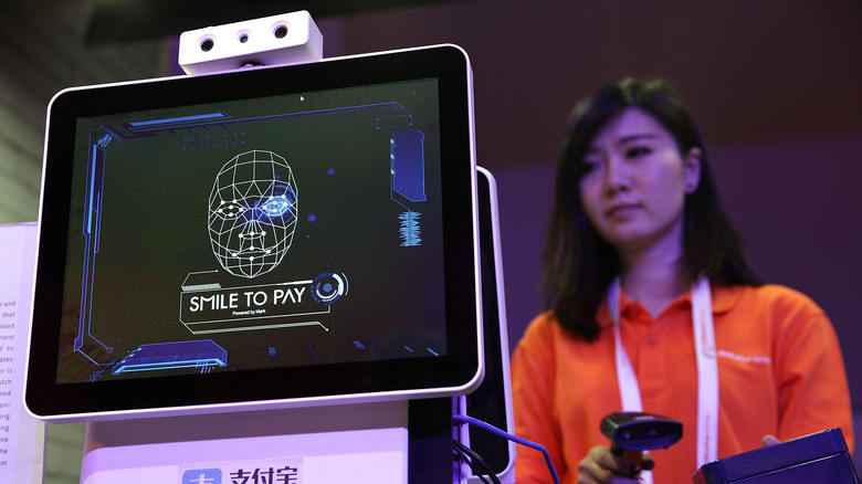 Demonstration of smile-to-pay checkout system on screen with woman holding scanner in background