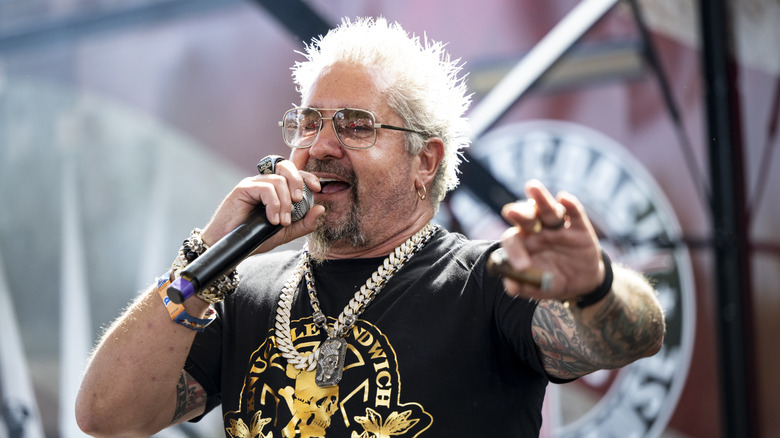 Guy Fieri speaks into a microphone, smiling