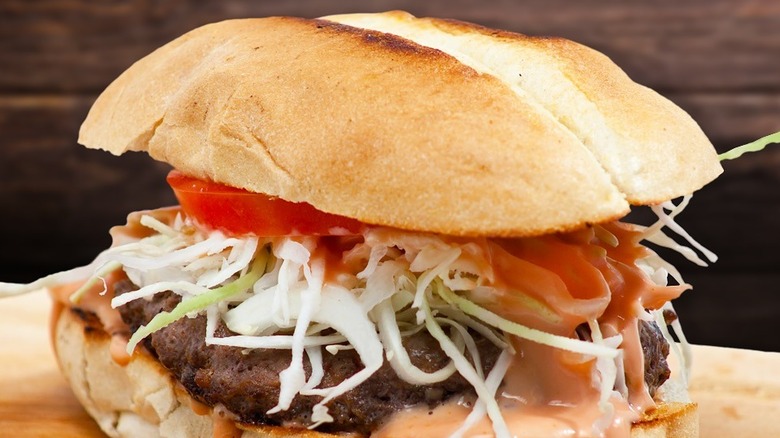 A dominican chimi featuring a patty, wilted cabbage, tomato, and pink sauce on a pan de agua