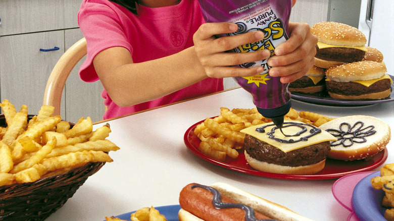 Kid applying purple color EZ Squirt ketchup on a hamburger