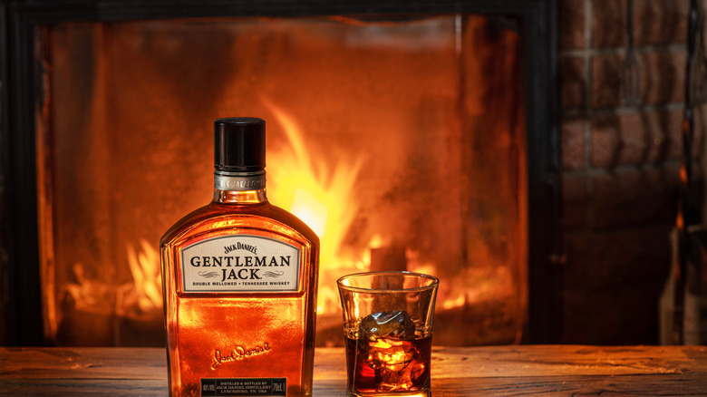 A bottle of Jack Daniel's Gentleman Jack Tennessee whiskey sits on a wooden table beside a large shot glass containing some of the spirit on the rocks. In the background, a fire burns in the hearth.
