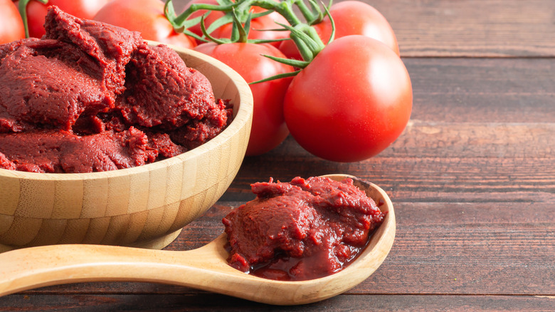 A wooden spoin and bowl of tomato paste; whole tomatoes are also in the frame.
