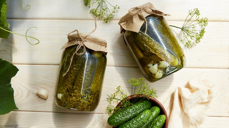 Two jars of cucumber pickles and a bowl of cucumbers on a wooden table.