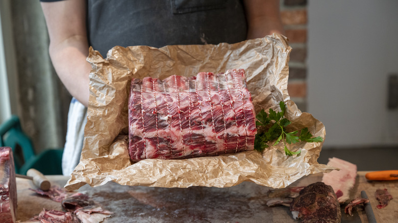 Butcher holding string-tied prime rib roast in butcher paper with herbs