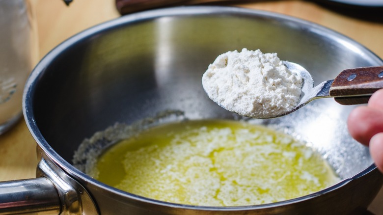 A spoonful of flour held over a stainless-steel pot with melted butter.