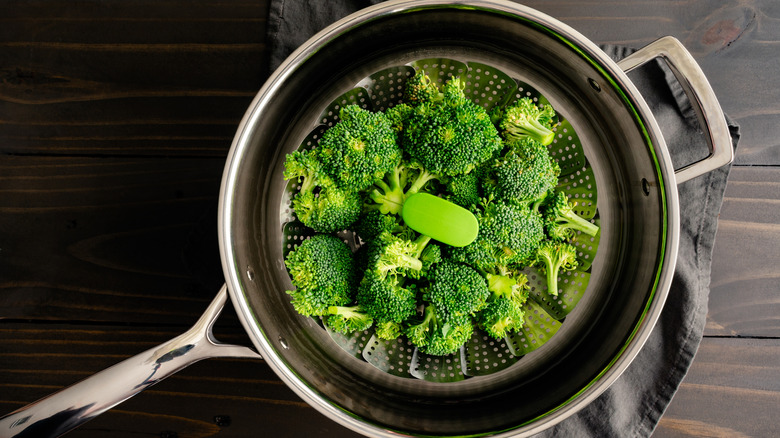 Raw broccoli florets in a foldable metal steaming basket placed in a pot with water