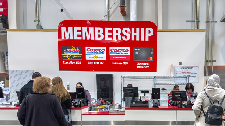 A Costco membership counter where customers are being assisted by employees, with a large sign displaying membership options