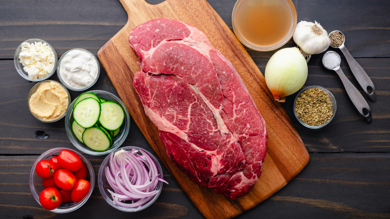 Various ingredients for pot roast surrounding a large slab of raw meat on cutting board