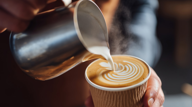 A person's hands are seen creating latte art in a cup with a chrome pitcher of steamed milk.
